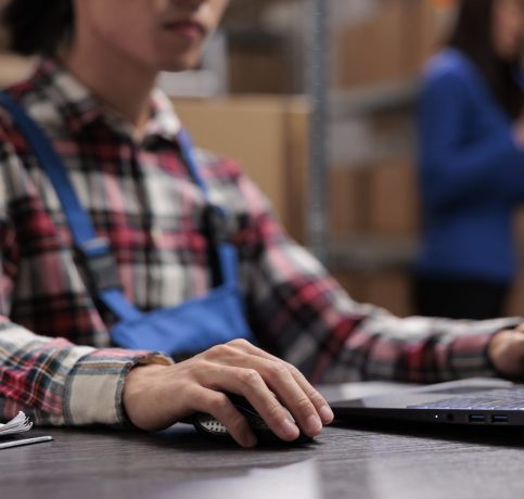 young-asian-logistics-manager-analyzing-goods-supply-laptop-warehouse-worker-doing-inventory-management-computer-while-sitting-desk-with-close-up-selective-focus-hand-min